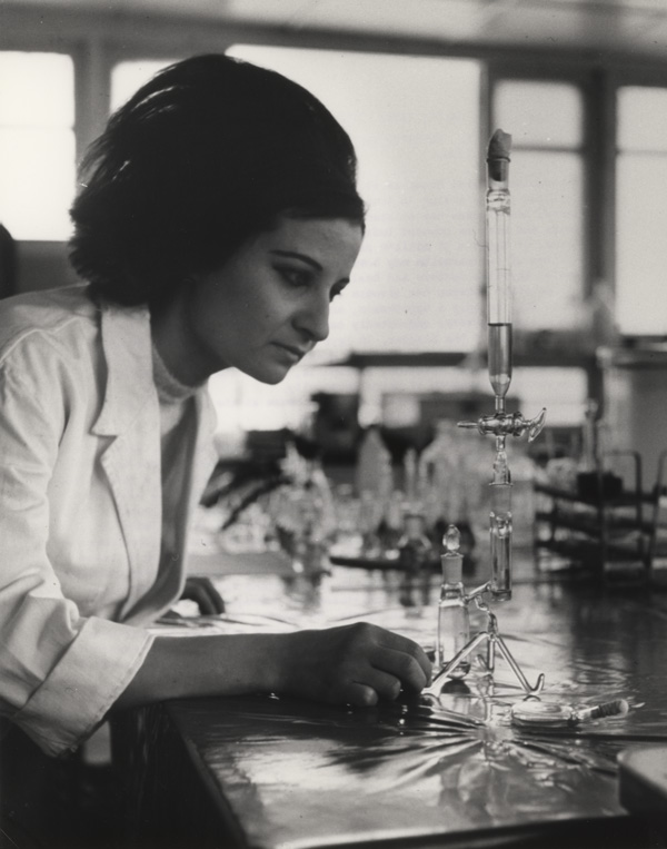 A woman leans toward a tall glass instrument on a laboratory desk.