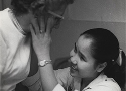 A woman stands and leans toward a seated smiling girl who is touching the woman’s right cheek.