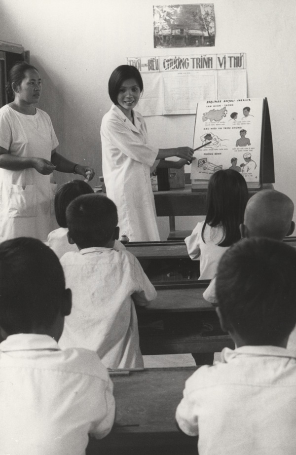 In front of a classroom, two women stand facing seated students as one points to a chart and the other stands on the side. 