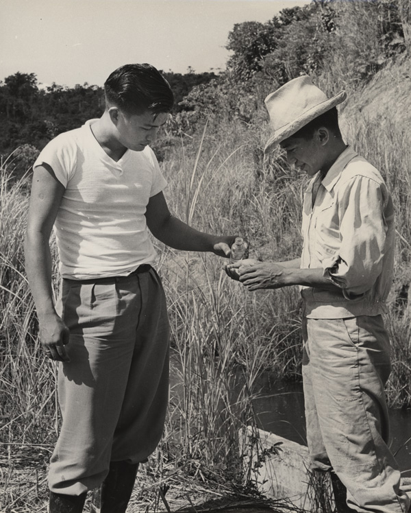 Outside, a man stands with his right palm open as another man holds a small bottle tilted over the palm. 