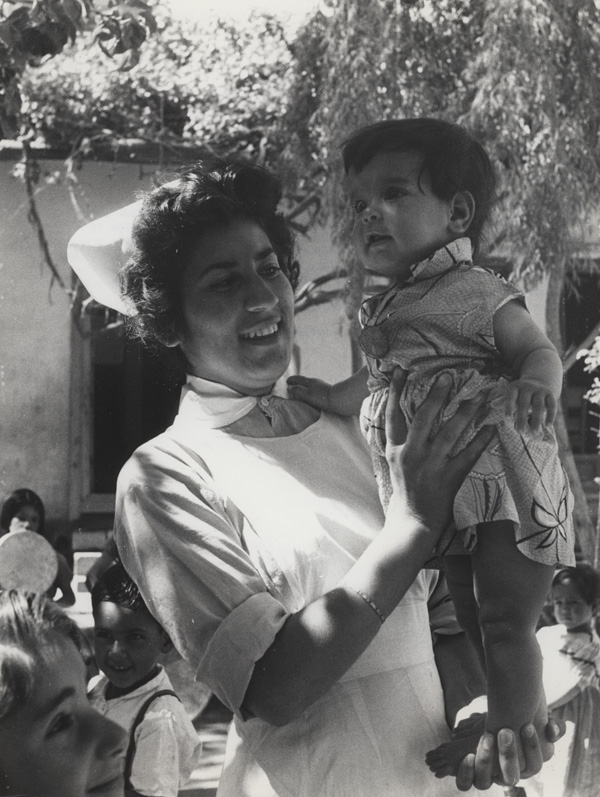 Outside, a woman in nurse’s uniform smiles while holding a child standing on her left palm.