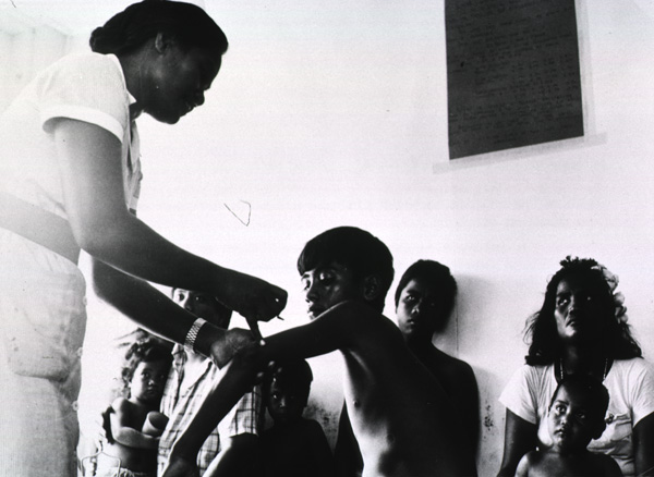 A nurse stands and gives an injection to a seated boy’s upper left arm as others look seated in the background.