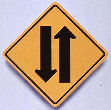 Yellow square road sign that has two vertical arrows pointing in opposite directions, above text