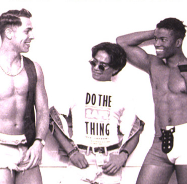 Black and white photograph of a multiracial group of men variously dressed waiting in line for a condom dispenser