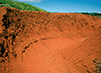 A depression of rust-colored earth, the result of soil erosion, is pictured. In the background are green grass and trees, and blue sky.