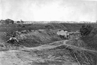 Black and white photograph of wooden barracks of Chimborazo Hospital, Richmond, Virginia in the distance and a man crouching down in the foreground. Courtesy Library of Congress.