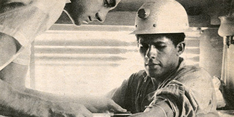 A white man standing leans down while taking blood from a seated Native American man in a hard hat.