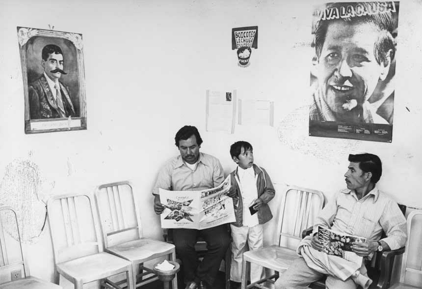 Two Latino men and one child in a clinic waiting room. The two men are seated and reading.