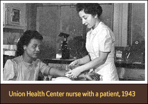 Standing White female nurse holds the arm of a seated African American woman.