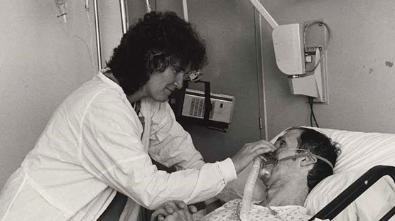 White female nurse smiles down as she places an oxygen mask on White male patient in bed.