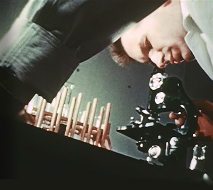 Man in lab coat leaning to look into a black microscope, with glass test tubs nearby on a table. 