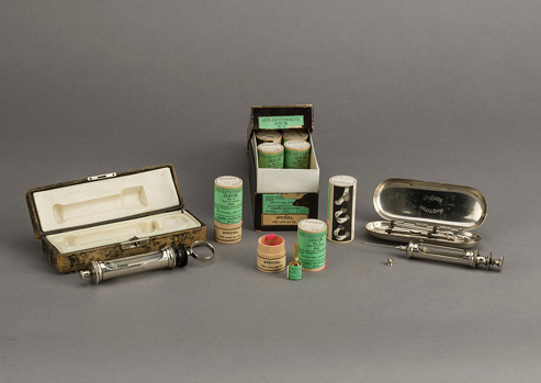 Box of wood canisters with green labels; small glass serum ampule is next to one open canister; two serum syringes with their cases are displayed on either side of box.