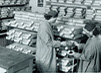 Two women in surgical gowns, masks, and head coverings in a room stacked with square ceramic penicillin vessels.