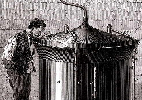 A man stands next to a large cylindrical beer vat and gazes into the vat through a small opening in the conical cover. 