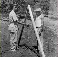 Two African men building wooden A-frame structures on a farm.