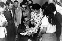 A Latino man sits and holds a child while a crowd stand around looking in.