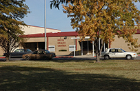 The entrance of a building with a street, parked cars, trees, and a lawn in front.