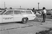 Two men help a patient on a gurney out of a car.