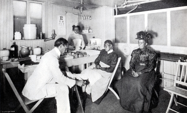 Physician seated in a chair tending to the leg of a seated patient with a nurse standing in the background.