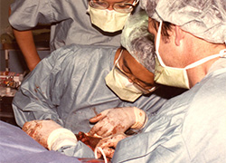 Three in surgical scrubs and masks in surgery: two next to the patient and one looking over a surgeon into the operation. 