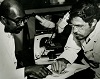 African American man and Hispanic man both look down and point at a book between them.