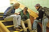 A White man and woman talk with a seated White man on a stairwell. 