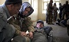 A White woman in military uniform tends to a Middle Eastern man in a stretcher as another man looks on.