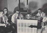 Four seated White men in lab coats look on as a White woman points towards a back lit board.