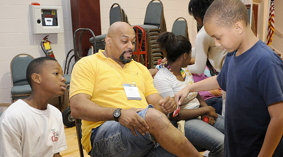 African American boy examines a seated African American man's knee as another boy looks on.