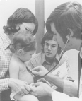 A National Health Service Corps physician listens to the heartbeat of an infant being held by its mother while the father watches in the background.