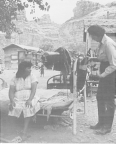 A woman sits on a bed that is outdoors. A health care worker stands at the foot of the bed with medications.