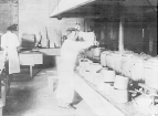 Interior view: a man stirs substance in huge pots lined in a row on a large stove; empty cans sit on a shelf in front of the pots; another man works in the background.