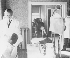 Black and white photograph from the examining into the waiting room of trachoma hospital in Rolla, Missouri. A physician looks at the eyes of a patient seated in a examination chair.