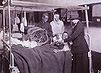 Photograph of two injured men in a hospital ward being handed cigarettes by a man and a woman.