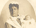 An African American female nurse in white holding a White infant, looking at the viewer.