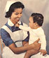 A Surinamese nurse in blue and white smiles at a Surinamese baby on her lap.