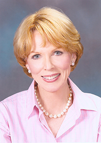 Dr. Bernadine Healy, a smiling White female in a pink collared shirt and pearls posing for her portrait.