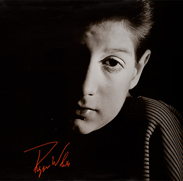 Black and white photograph of a teenage boy (Ryan White), looking at the viewer with half his face covered in shadow, White’s signature is included