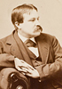 A man with a mustache wearing suit and leaning on a chair arm in profile facing the right.