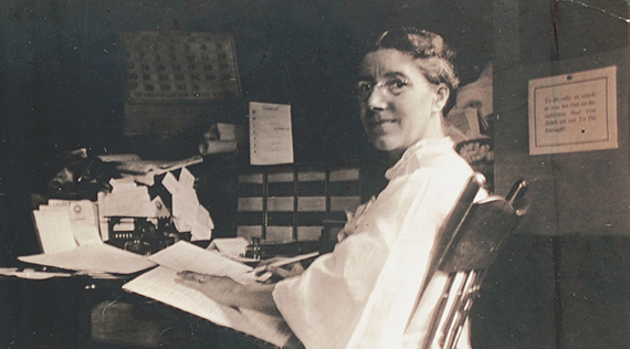 A woman at seated at a desk, turning towards her left to look at viewer.