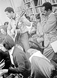 A roomful of people listens to an African American man sing and play guitar