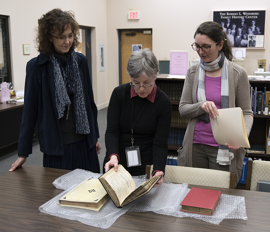 Three women examine books on a table.