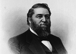 A formal photograph of a full bearded man in a suit with a printed autograph at the bottom.